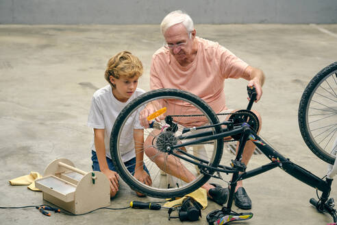 Konzentrierter älterer Mann mit Enkel, der ein Fahrradrad repariert, während er auf dem Betonboden sitzt - ADSF46820