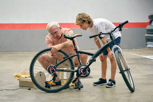 Ganzkörperaufnahme eines ernsten Großvaters, der die Reifen eines Fahrrads aufpumpt, in der Nähe eines Enkels, der das Rad mit Aufmerksamkeit betrachtet - ADSF46821