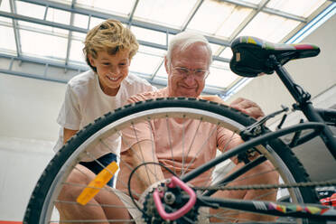 Positiver Großvater mit grauem Haar und Brille repariert Fahrrad mit Unterstützung eines Jungen, der in der Nähe steht - ADSF46823