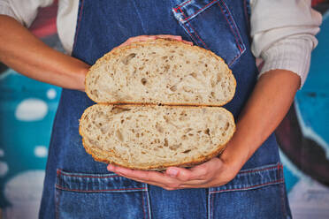 Fotoausschnitt einer weiblichen Hand, die ein in der Mitte gebrochenes Brot mit einer blauen Küchenschürze hält. - ADSF46828