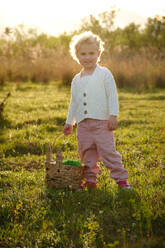 Full body content little girl in stylish clothes with wicker basket standing on abundant grassy meadow on sunny day and looking at camera with pretty smile - ADSF46834