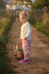 Side view adorable little girl in stylish clothes carrying wicker basket and standing on narrow rural road in verdant nature - ADSF46836