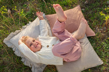 From above smiling little girl in stylish wear lying with legs raised on soft pillows on grassy meadow in summer countryside - ADSF46837