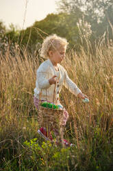 Side view full body cute little girl in stylish wear collecting colorful Easter eggs into wicker basket in lush countryside on sunny spring day - ADSF46839