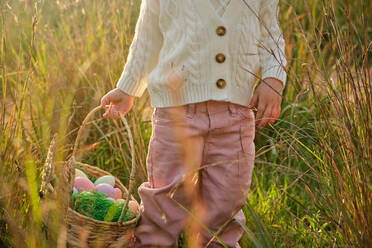 Crop unerkennbar Mädchen in weißem Pullover trägt bunte Ostereier in Weidenkorb, während stehend auf grasbewachsenen Rasen in sonnigen Landschaft - ADSF46841