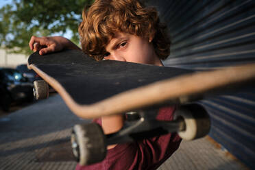 Focused boy with curly hair examining skateboard while standing on sidewalk in summer on city street - ADSF46845