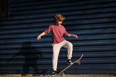 In voller Länge kleine Skater in Freizeitkleidung tun Trick auf Skateboard gegen dunkelblaue Wand auf der Straße der Stadt - ADSF46846