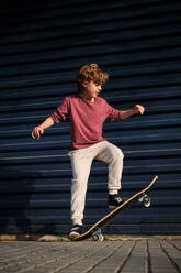 Ganzkörper-Junge in Freizeitkleidung macht Stunt auf dem Skateboard auf dem Bürgersteig in der Nähe von Metall Gebäude Wand - ADSF46847