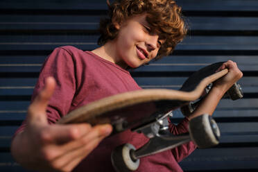 Von unten positive kleine Skater lächelnd und Überprüfung Skateboard in der Nähe von dunkelblauen Wand in der Stadt stehen - ADSF46850