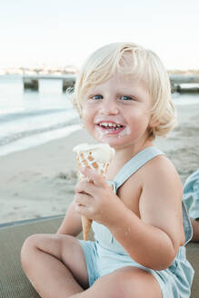 Niedlicher kleiner Junge in Freizeitkleidung lächelt und schaut in die Kamera, während er am Strand sitzt und Eis isst - ADSF46858