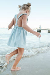 Side view of anonymous little barefoot girl in summer dress running on wet beach washed by foamy sea - ADSF46860