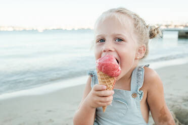 Bezauberndes kleines Mädchen isst süßes Eis und schaut in die Kamera, während es am Sandstrand in der Nähe des wogenden Meeres sitzt - ADSF46862