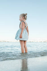 From below side view of happy little girl in summer dress standing in shallow sea on beach and looking away against cloudless sky - ADSF46863