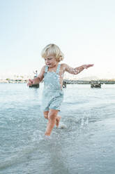 Full body of cute little toddler playing and running in rippling sea on sunny summer day - ADSF46866