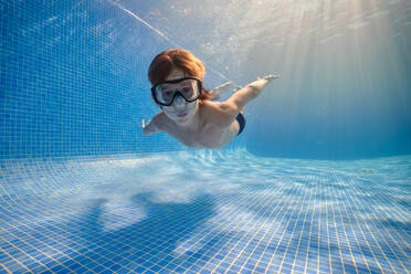 Boy in goggles and shorts swimming in clear transparent water above sunlight shining through water and looking at camera - ADSF46873