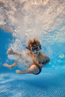 Ganzer Körper eines barfuß gehenden Jungen mit Schwimmbrille, der im klaren, transparenten Wasser eines Schwimmbads mit Blasen springt - ADSF46875