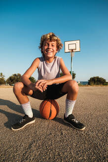 Ganzkörperaufnahme eines fröhlichen Jungen in Sportkleidung, der auf einem Basketball auf einem Spielplatz sitzt und in die Kamera schaut - ADSF46879