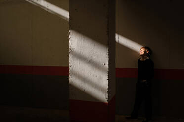 Ganzkörperaufnahme eines niedlichen Teenagers, der an der Wand hinter einer Betonsäule steht und einen Sonnenstrahl im Gesicht hat - ADSF46886
