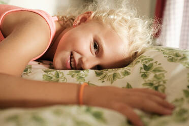 Seitenansicht eines lächelnden blonden Mädchens, das auf dem Bett liegt und in die Kamera schaut, während es sich im Schlafzimmer entspannt - ADSF46893