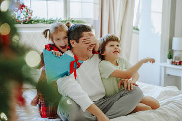 Tochter hält Weihnachtsgeschenk und bedeckt die Augen des Vaters im Bett - NLAF00111