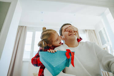 Lächelnde Tochter, die ein Weihnachtsgeschenk hält und dem Vater zu Hause die Augen zuhält - NLAF00112