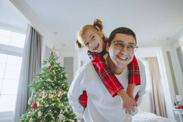 Lächelnder Vater, der seine Tochter in der Nähe des Weihnachtsbaums zu Hause huckepack nimmt - NLAF00124