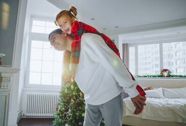 Glücklicher Vater, der seine Tochter in der Nähe des Weihnachtsbaums zu Hause huckepack nimmt - NLAF00125