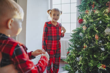 Lächelnde Schwester betrachtet Bruder in der Nähe von Weihnachtsbaum zu Hause - NLAF00132