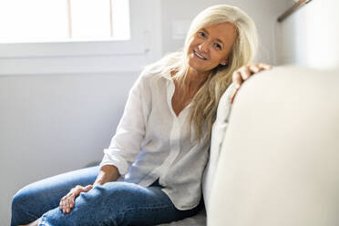 Lächelnde reife Frau sitzt auf dem Sofa zu Hause - DLTSF03620