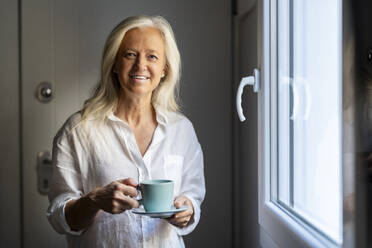 Lächelnde reife Frau stehend mit Kaffeetasse zu Hause - DLTSF03628