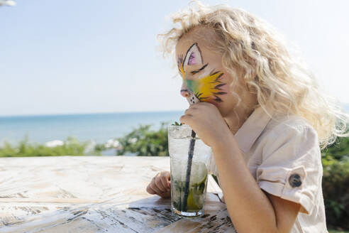 Blondes Mädchen mit Gesichtsbemalung trinkt Mojito am Tisch - SIF00909