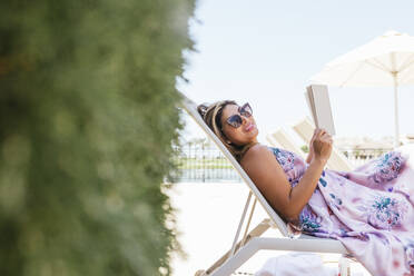 Glückliche Frau mit Sonnenbrille und entspannt mit Buch auf einem Liegestuhl - DSIF00709