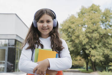 Lächelndes Schulmädchen mit drahtlosen Kopfhörern und Büchern in der Hand - OSF02132