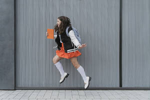 Smiling schoolgirl holding book and jumping in front of wall - OSF02136