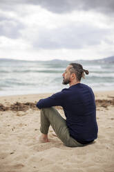 Thoughtful man sitting on sand at beach - JOSEF21039