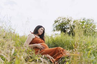 Schwangere Frau sitzt im Gras unter dem Himmel - NDEF01164