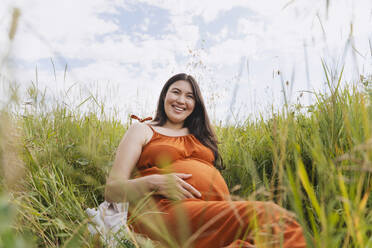 Glückliche schwangere Frau sitzt im Gras unter dem Himmel - NDEF01165