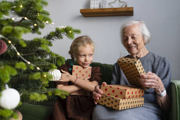 Glückliche Enkelin mit Großmutter, die Geschenke am Weihnachtsbaum zu Hause hält - SVKF01610