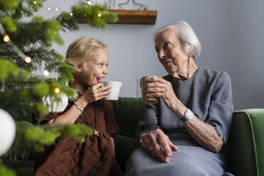 Glückliches Mädchen, das mit seiner Großmutter heiße Schokolade trinkt und seine Freizeit in der Nähe des Weihnachtsbaums zu Hause verbringt - SVKF01612