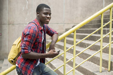 Glücklicher junger Mann, der Musik hört und sein Smartphone hält und sich an ein Geländer lehnt - IKF01231