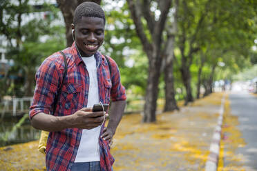 Smiling young man wearing in-ear headphones listening to music and using smart phone - IKF01235