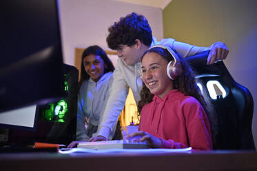 Lächelndes junges Teenager-Mädchen mit Kopfhörern, das auf den Bildschirm eines Monitors schaut, während es mit einem Spiele-PC am Tisch sitzt und auf seine multirassischen Freunde im Zimmer schaut - ADSF46946