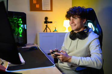 Fokussierter lächelnder junger Mann, der auf den Bildschirm eines Computers schaut, während er an einem Tisch mit einem Spiele-PC sitzt und allein mit einem Joystick im Zimmer Videospiele spielt - ADSF46952