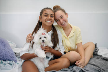 Lächelnde diverse Teenager-Freundinnen sitzen auf dem Bett und umarmen einen Hund, während sie in die Kamera schauen - ADSF46956