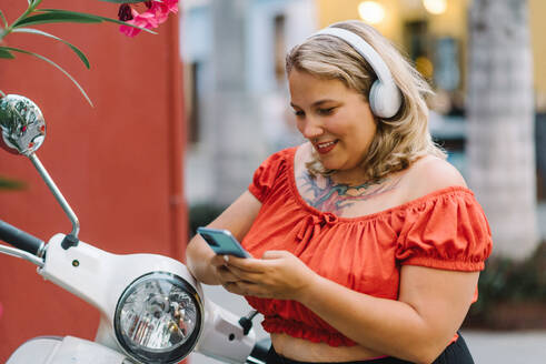 Seitenansicht von plump junge Frau in Kopfhörer lächelnd und Blick auf Smartphone, während stehend mit modernen Roller auf Stadtstraße mit Unschärfe Hintergrund - ADSF46957