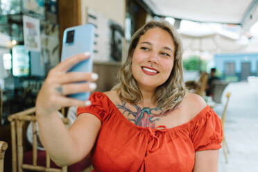 Portrait of plump young female in casual clothes and tattoo smiling while taking selfie on smartphone outdoors in cafe on blur background in daylight - ADSF46963