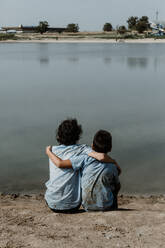 Back view of unrecognizable boys in dirty school uniform and curly hair sitting on shore of lake with hands over shoulder while enjoying summer day - ADSF46970