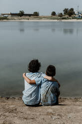 Rückenansicht eines nicht erkennbaren Jungen in schmutziger Schuluniform und lockigem Haar, der am Ufer eines Sees sitzt und die Hände über die Schulter legt, während er den Sommertag genießt - ADSF46970