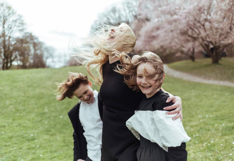 Glückliche Frau und Kinder in schwarzen Outfits stehen auf grünem Gras in der Nähe von blühenden Kirsche - ADSF46975