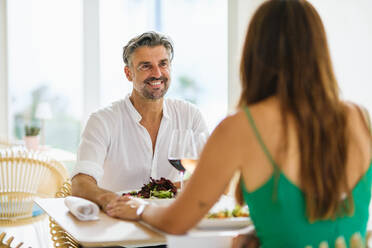 Verliebtes Paar in eleganter Kleidung, das sich lächelnd anschaut und die Hände hält, während es am Tisch mit leckeren Salaten und Wein bei einem Date im Restaurant auf der Straße sitzt - ADSF46988